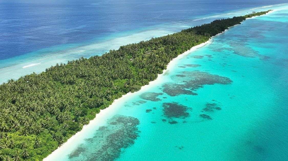 Una vista aerea di una lunga e stretta isola tropicale coperta di palme, con una spiaggia di sabbia bianca, una barriera corallina turchese e un oceano blu profondo.
