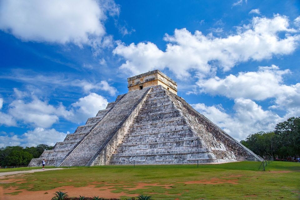 Una grande piramide a gradoni di pietra si erge su un campo erboso sotto un cielo azzurro brillante con nuvole bianche sparse.