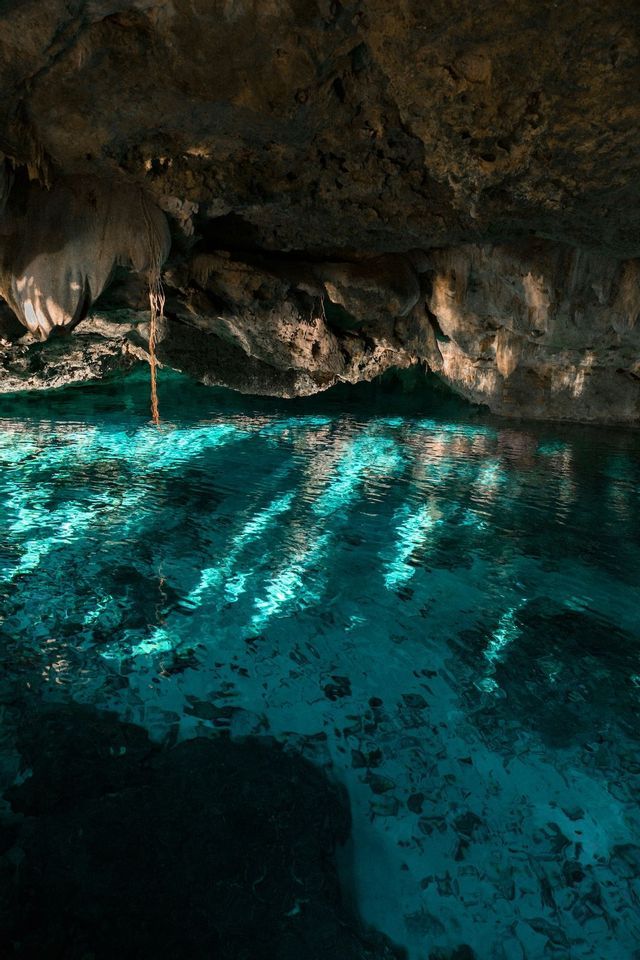 La luce del sole si riversa in una grotta buia e rocciosa, creando giochi di luce sull'acqua turchese e cristallina sottostante.