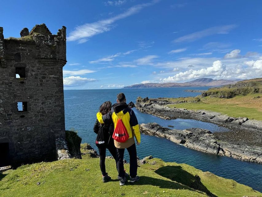 Two people on a WeRoad group trip stand on a grassy cliff beside a stone ruin, looking out at the sea and rocky coastline.