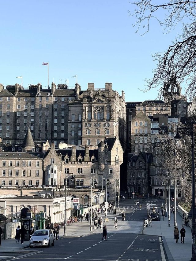 A sunlit city street with pedestrians walking alongside historic stone buildings under a clear blue sky.