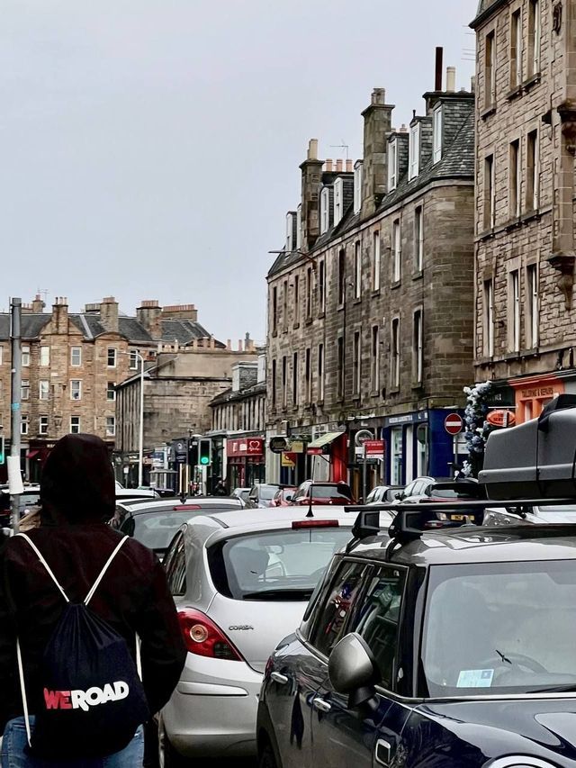 Une personne de dos, portant un sac à dos WeRoad, marche dans une rue de ville bordée de voitures et de bâtiments en pierre.