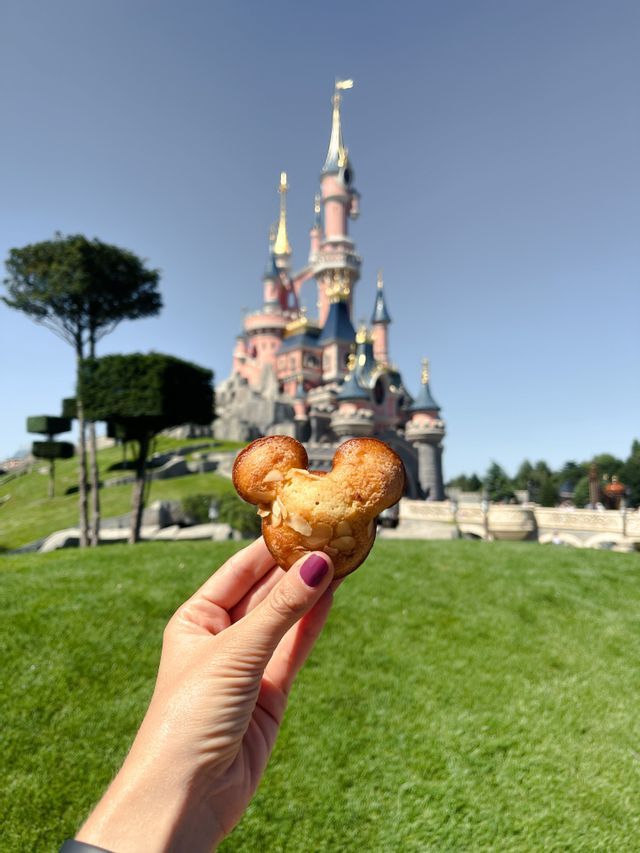 Eine Hand hält ein Micky-Maus-förmiges Gebäck vor einem großen, rosafarbenen Märchenschloss auf einem Rasen.
