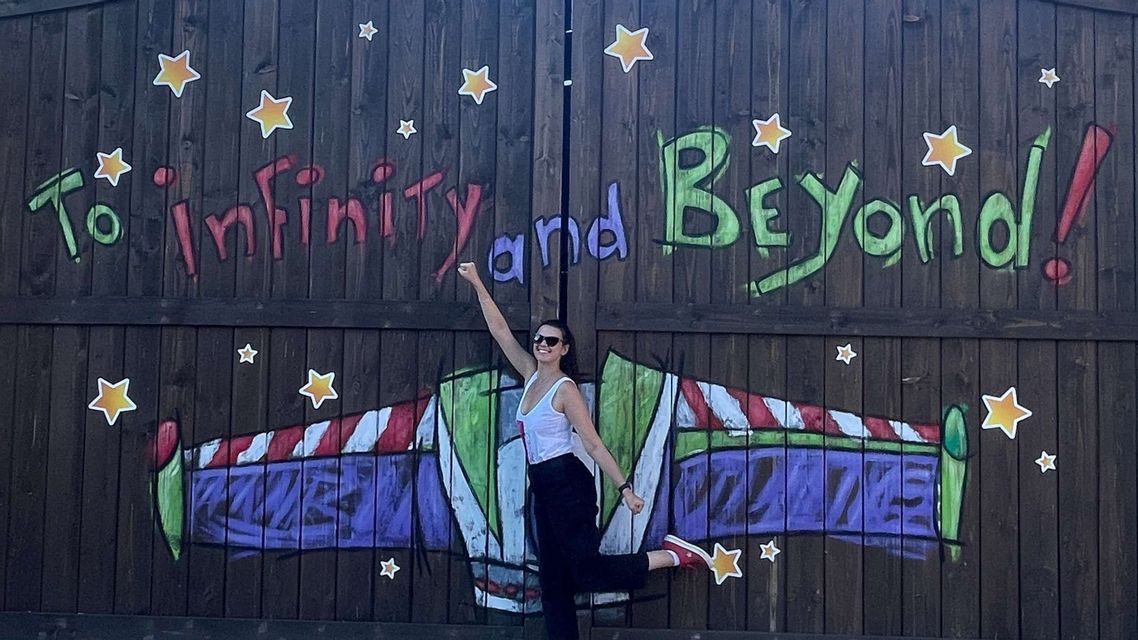 Eine lächelnde Frau posiert verspielt vor einer Holzwand mit dem Graffiti 'Ins Unendliche und darüber hinaus!'