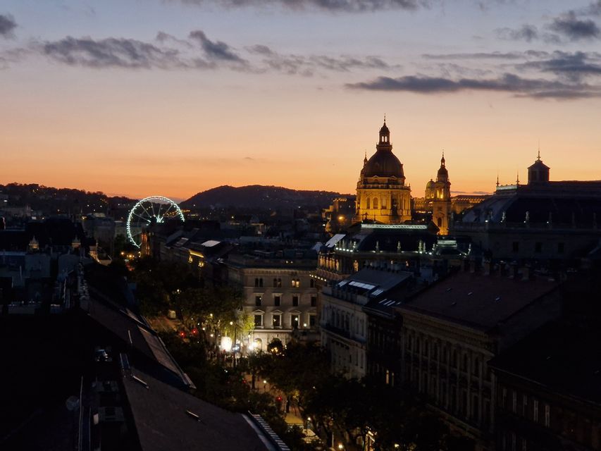 Eine Stadtansicht bei Sonnenuntergang mit einer beleuchteten Basilika und einem großen Riesenrad vor einem orange-lila Himmel.