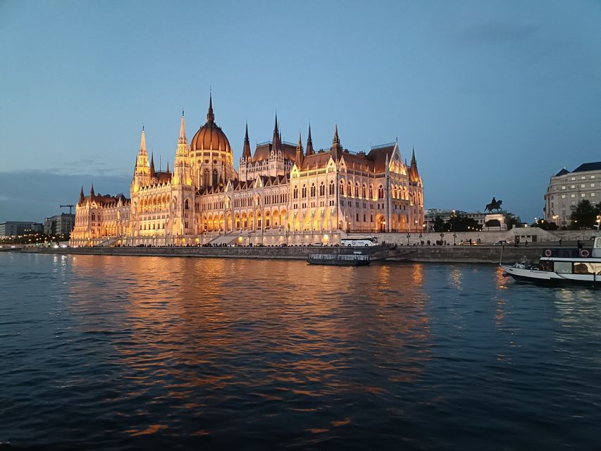 Ein prunkvolles, beleuchtetes Parlamentsgebäude an einem Flussufer in der Abenddämmerung, dessen Lichter sich im Wasser spiegeln.
