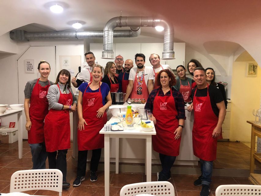 Un gruppo WeRoad sorride per una foto durante una lezione di cucina, tutti indossano grembiuli rossi coordinati in una cucina.
