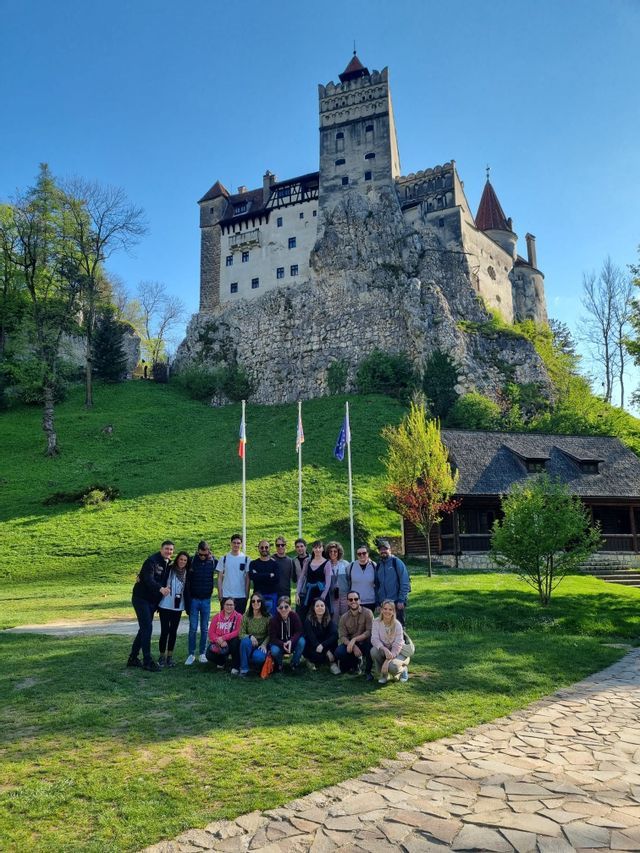 Un gruppo WeRoad in viaggio posa per una foto in una giornata di sole davanti a un castello storico, arroccato su una verde collina rocciosa.