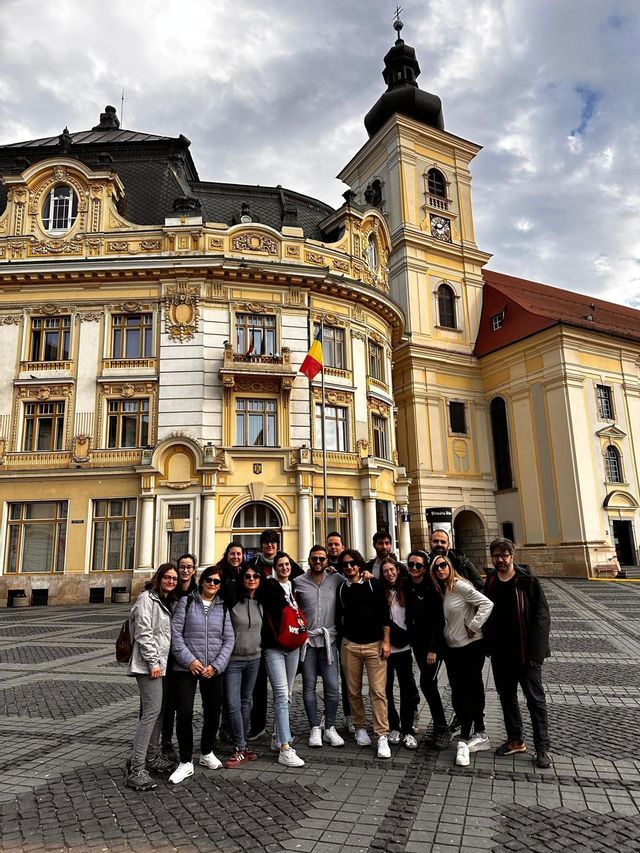 A WeRoad group trip poses for a photo in a cobblestone square in front of an ornate yellow building with a clock tower.