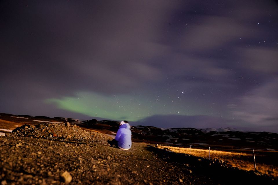 Una persona con giacca blu siede su un pendio roccioso di notte, osservando l'aurora boreale verde in un cielo stellato.