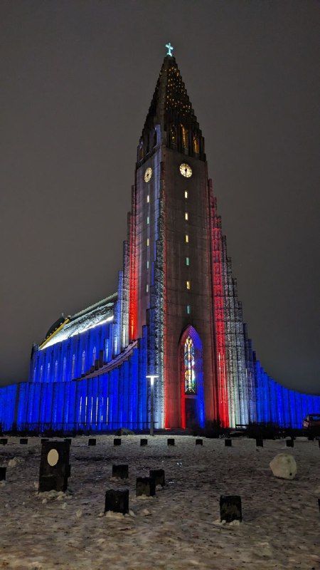 Una chiesa alta e moderna è illuminata di notte con luci blu, bianche e rosse, con neve sul terreno in primo piano.