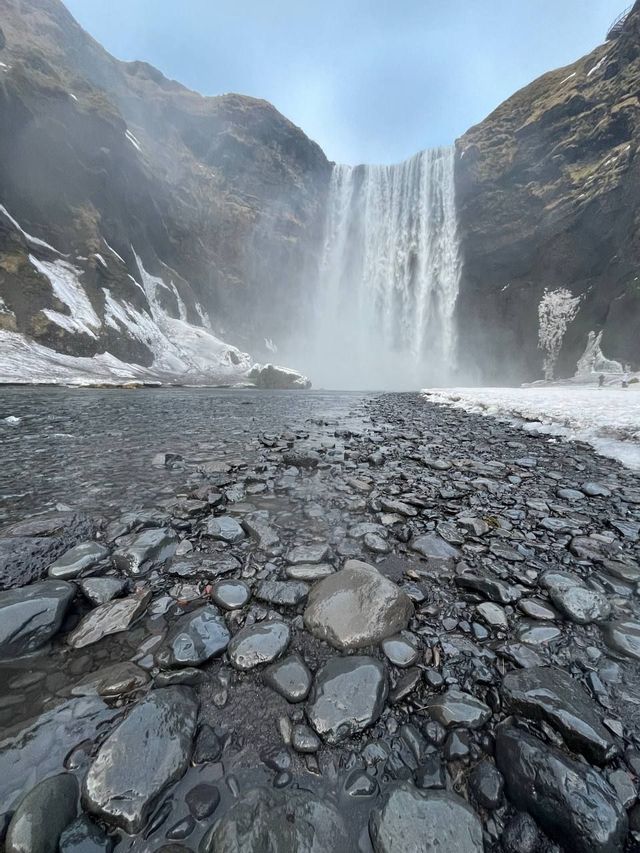Una grande cascata ripresa dal basso, che si getta tra scogliere innevate, con un letto di fiume roccioso e bagnato in primo piano.