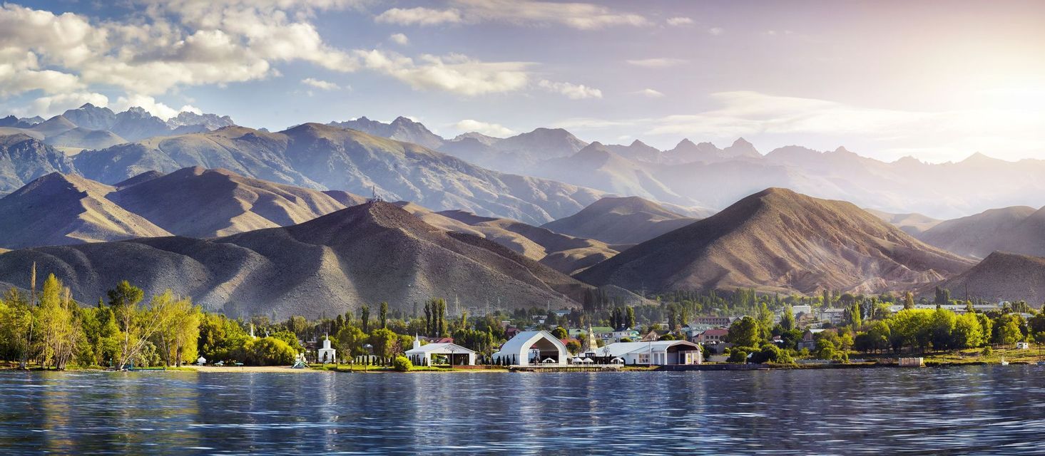Una cittadina sul lago con alberi verdi ed edifici bianchi si adagia ai piedi di grandi montagne aride sotto un cielo luminoso.