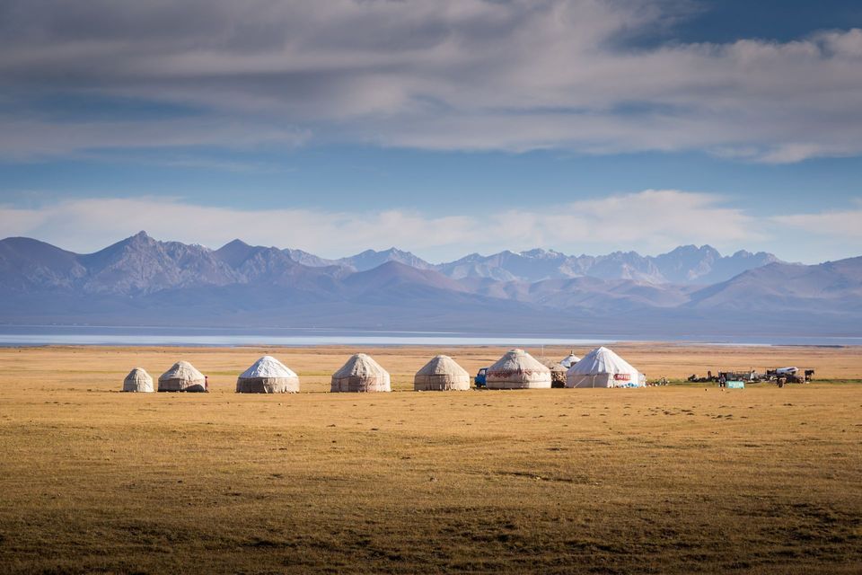 A row of yurts on a grassy plain in front of a lake and a distant mountain range.