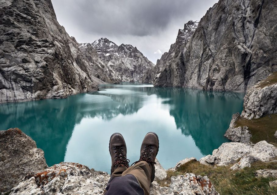 Vue à la première personne de chaussures de randonnée sur une corniche rocheuse, surplombant un lac turquoise calme entouré de montagnes abruptes sous un ciel nuageux.