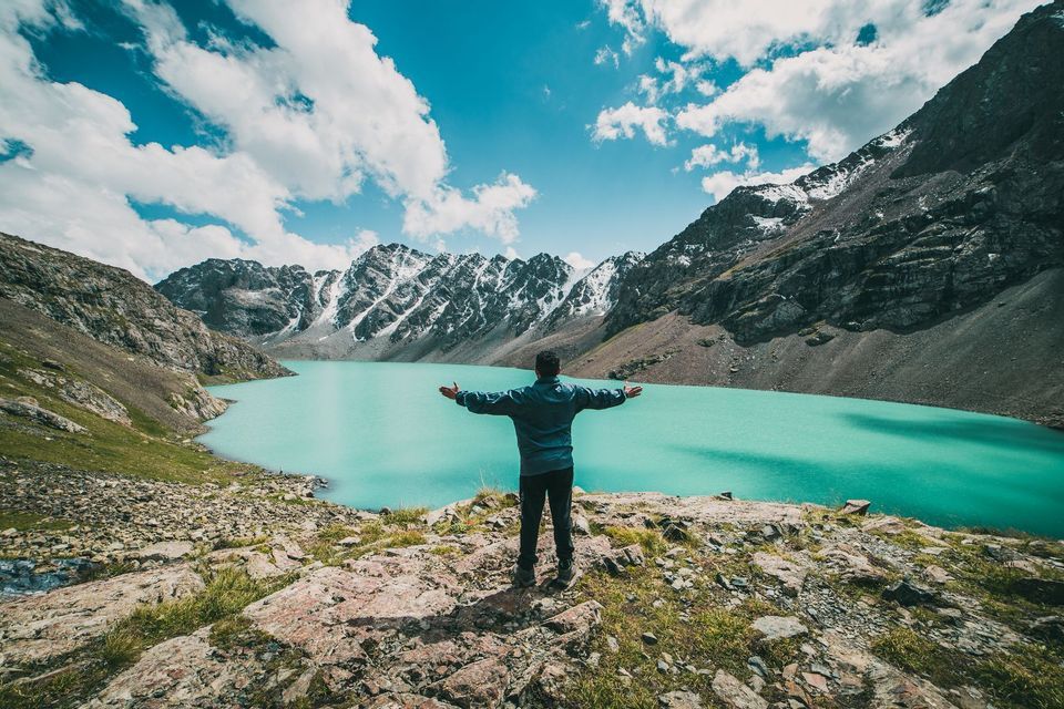 Una persona a braccia aperte si erge su uno sperone roccioso, ammirando un lago alpino turchese circondato da montagne innevate.