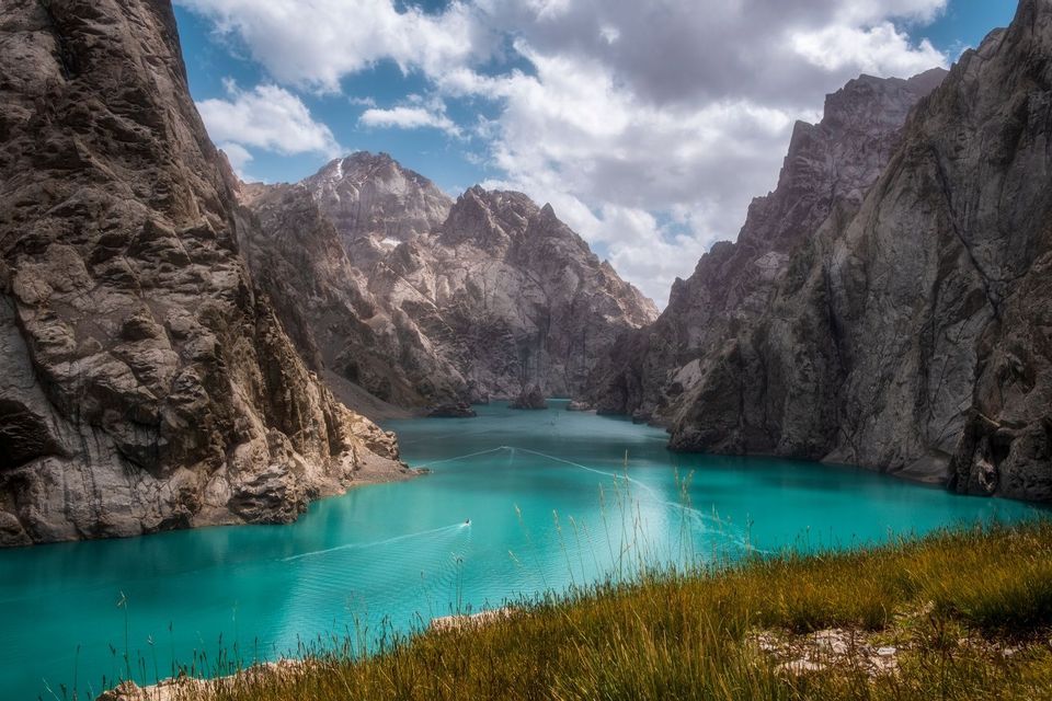 Una piccola barca lascia una scia su un lago turchese circondato da ripide montagne rocciose sotto un cielo nuvoloso.