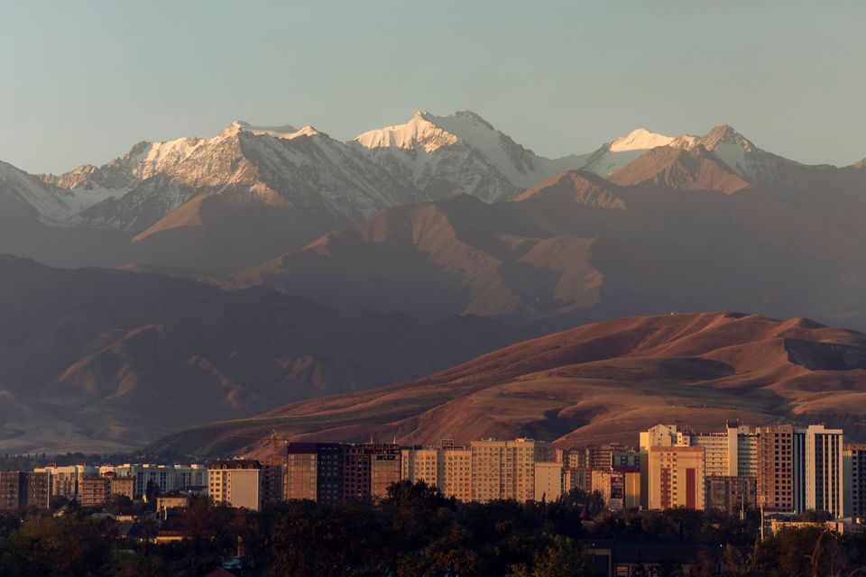 Uno skyline di edifici cittadini si erge ai piedi di dolci colline, con una grande catena montuosa innevata sullo sfondo.