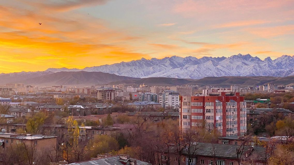 Un paysage urbain avec des bâtiments nichés devant une chaîne de montagnes enneigées sous un ciel de coucher de soleil doré.