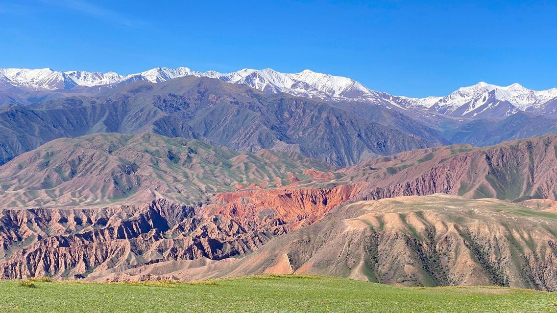 Un paysage étagé, avec des collines verdoyantes, des formations rocheuses rouges et une chaîne de montagnes enneigée au loin, sous un ciel dégagé.
