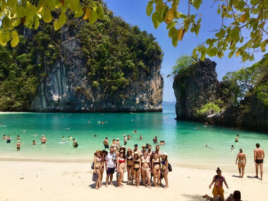 Un grupo de WeRoad posando en una playa de arena en una bahía turquesa, con grandes acantilados boscosos al fondo.