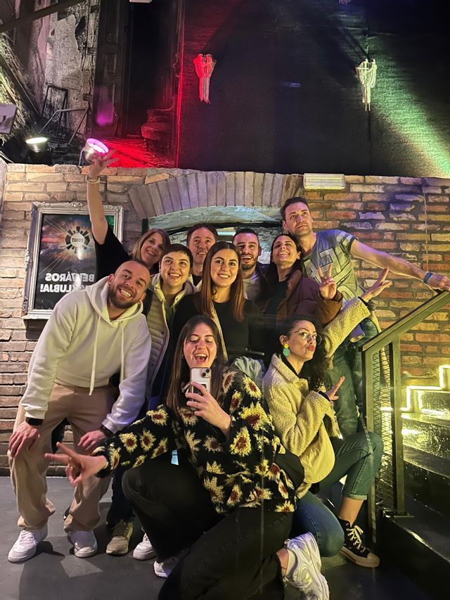 Un grupo de WeRoad sonríe y posa para una selfie grupal en una escalera dentro de un local con paredes de ladrillo y luces de colores.