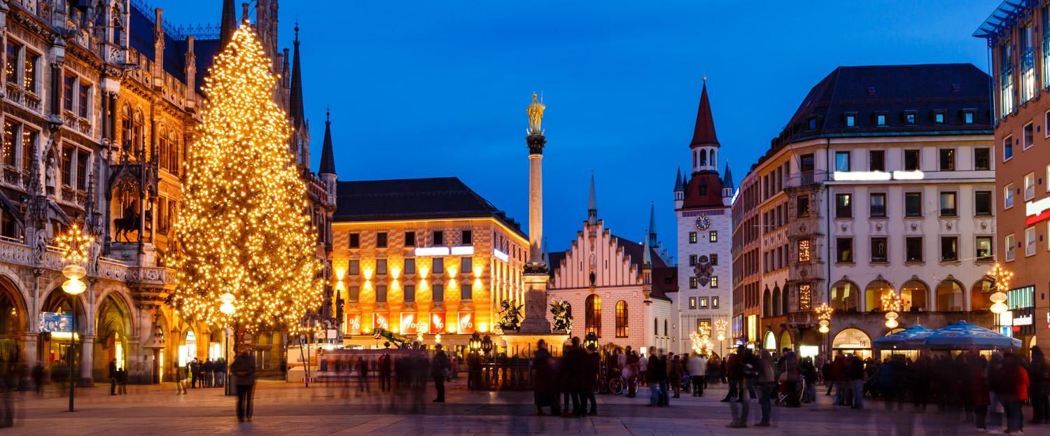 Un albero di Natale illuminato ed edifici storici si ergono su una piazza cittadina affollata al crepuscolo, con persone riprese con effetto mosso.