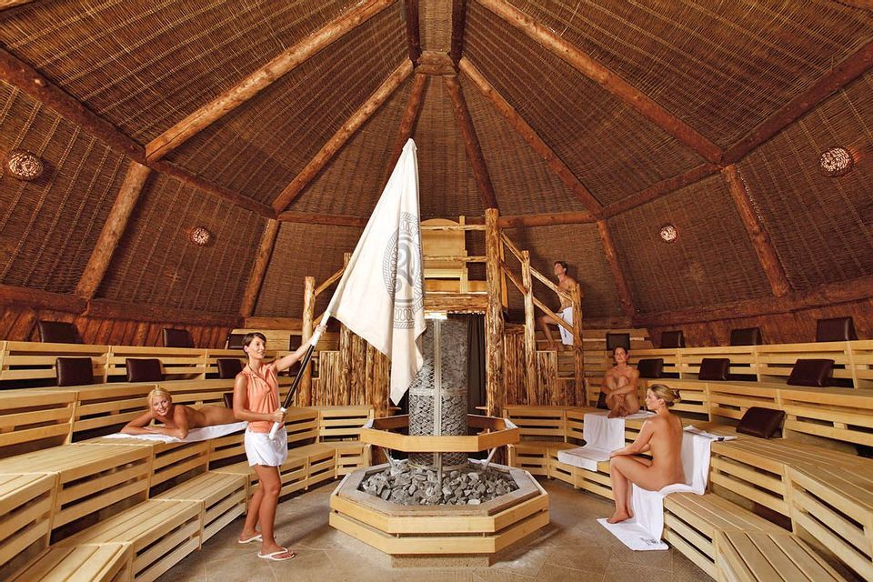 Una donna sventola una grande bandiera per far circolare l'aria tra le persone che si rilassano su panche di legno all'interno di una grande sauna circolare.