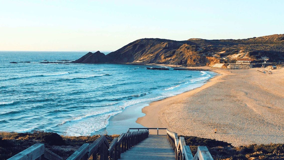 Vista da una scala di legno che si affaccia su una spiaggia sabbiosa con onde blu dell'oceano e una grande scogliera rocciosa.