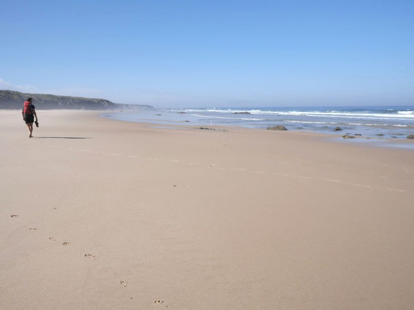 Una persona con un grande zaino cammina a piedi nudi su un'ampia spiaggia sabbiosa, lasciandosi dietro delle impronte.