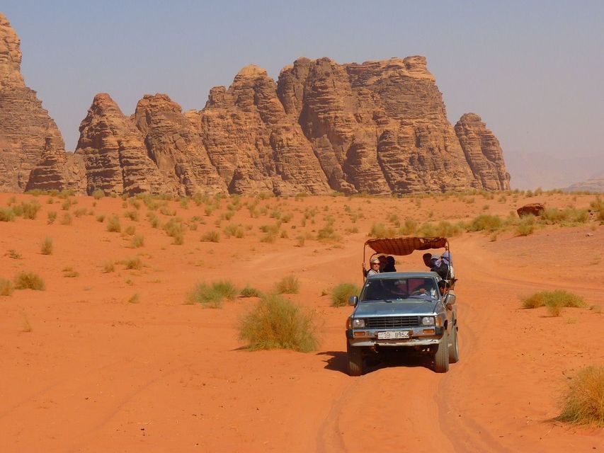 Un viaggio di gruppo WeRoad a bordo di un camion scoperto che attraversa un deserto di sabbia rossa con grandi formazioni rocciose sullo sfondo.