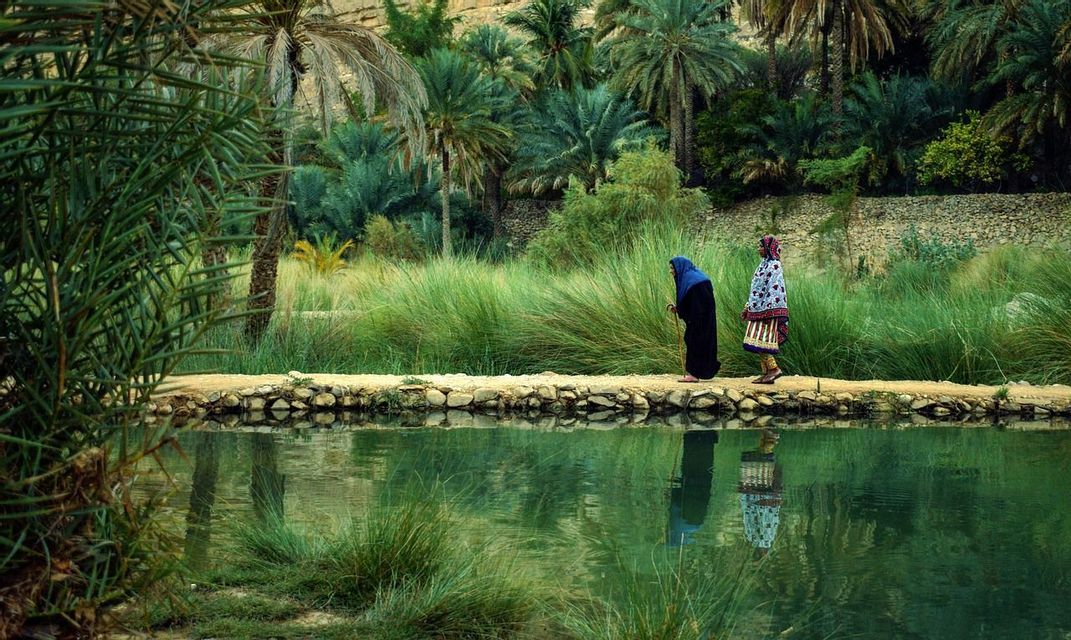 Due donne in abiti tradizionali camminano lungo un sentiero di pietra accanto a uno specchio d'acqua in una lussureggiante oasi verde con palme.