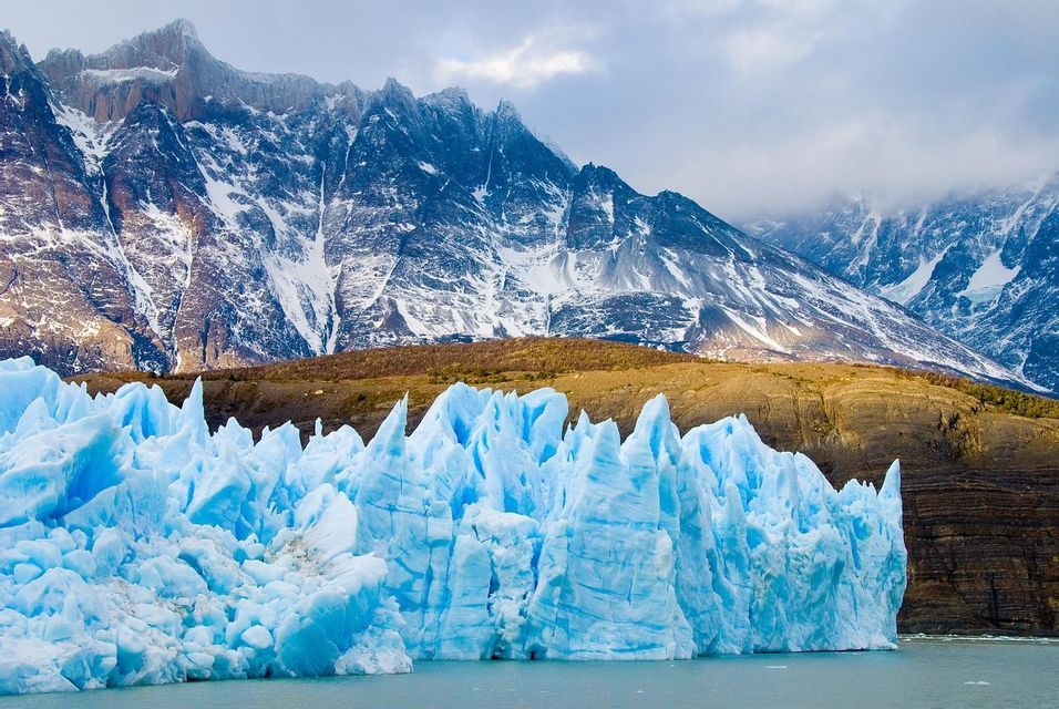 Un ghiacciaio azzurro brillante con guglie frastagliate galleggia nell'acqua di fronte a una catena di alte montagne innevate.