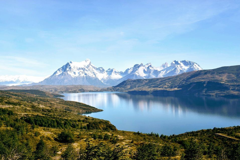 Un lago calmo e blu riflette una catena montuosa innevata e distante sotto un cielo sereno, con verdi colline in primo piano.