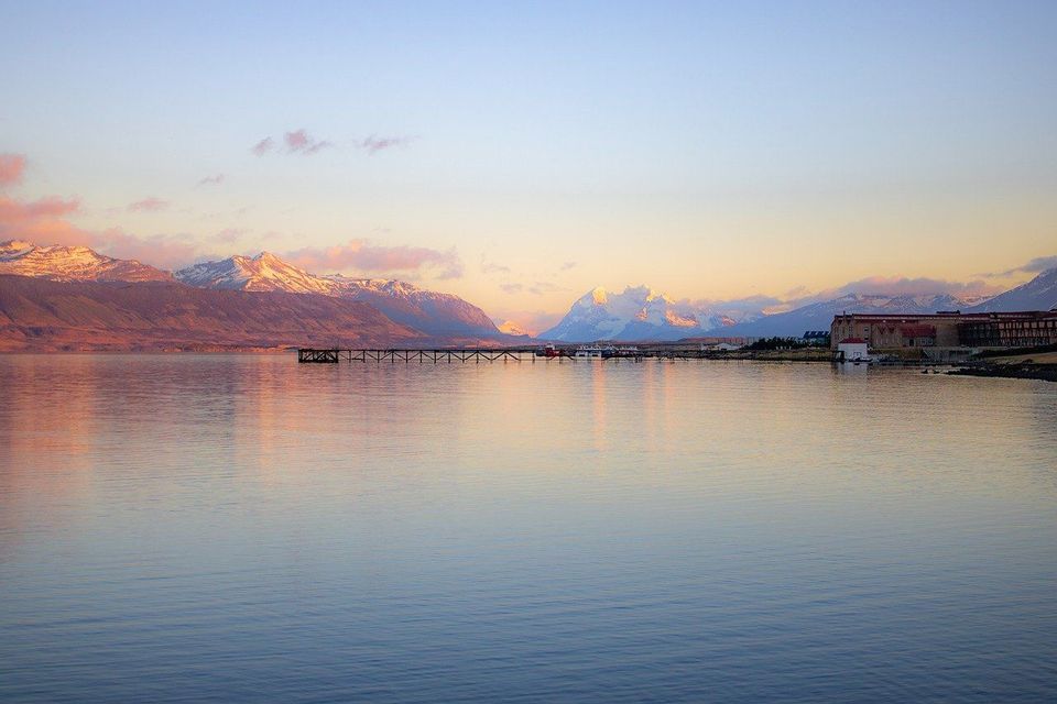 Un lago calmo riflette un cielo colorato al tramonto con montagne innevate sullo sfondo e un pontile sulla riva.