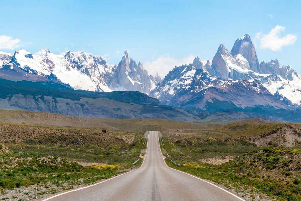 Una strada asfaltata vuota si estende attraverso un paesaggio verde verso una lontana catena montuosa innevata sotto un cielo blu.