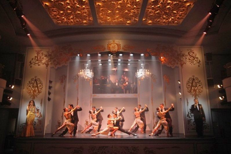 Coppie che ballano il tango su un sontuoso palcoscenico teatrale con cantanti e una band dal vivo sullo sfondo.