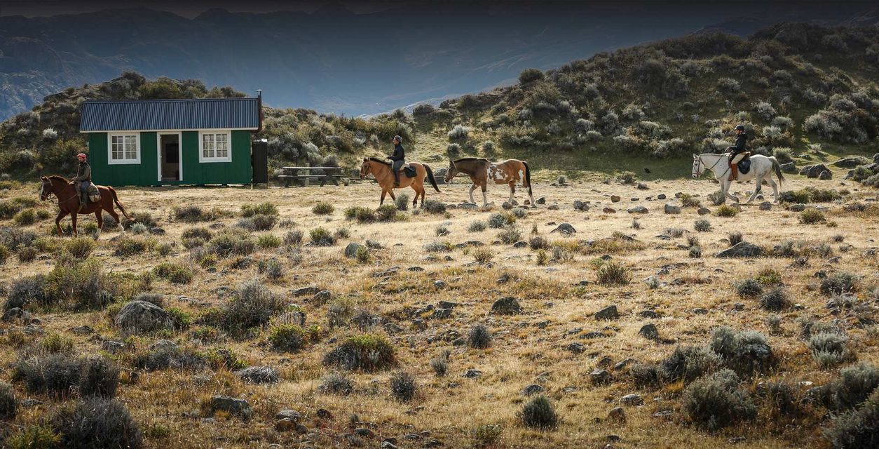 Un viaggio di gruppo WeRoad a cavallo attraversa un paesaggio arido e collinare, passando accanto a una piccola cabina verde sotto un cielo ampio.