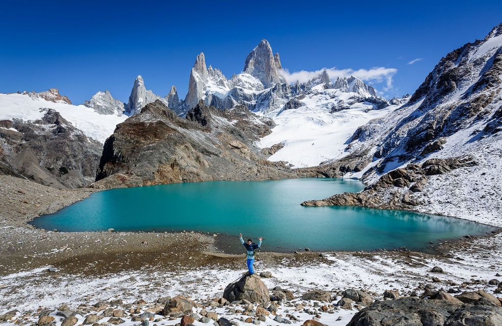 Una persona con le braccia alzate si erge su una roccia accanto a un lago alpino turchese, con montagne frastagliate e innevate sullo sfondo.
