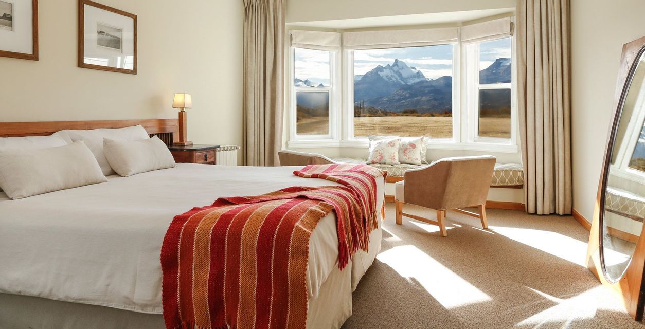 Una camera da letto illuminata dal sole con un letto fatto e un plaid a righe, dotata di una finestra a bovindo con vista su montagne innevate.