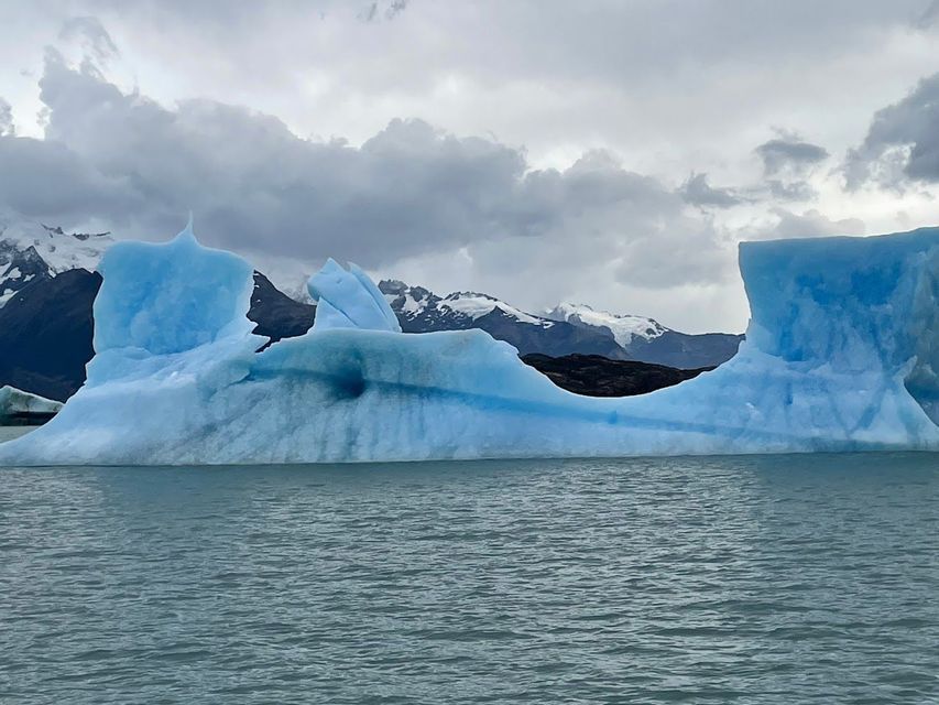 Un grande iceberg blu galleggia sull'acqua di fronte a montagne innevate sotto un cielo nuvoloso.