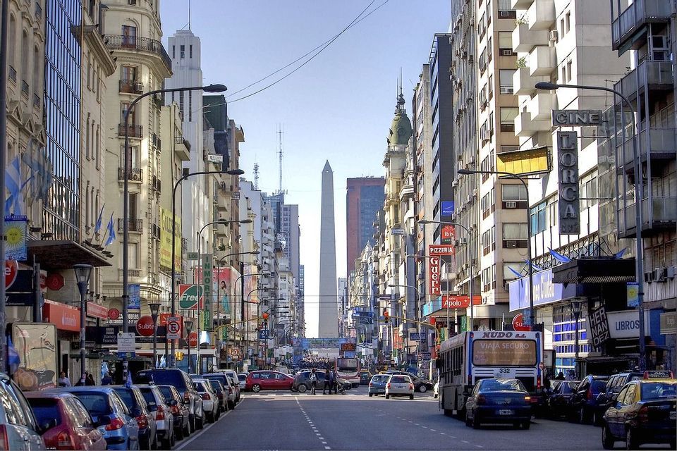 Un'ampia strada cittadina fiancheggiata da alti edifici conduce verso un grande obelisco bianco, con auto e un autobus sulla strada.