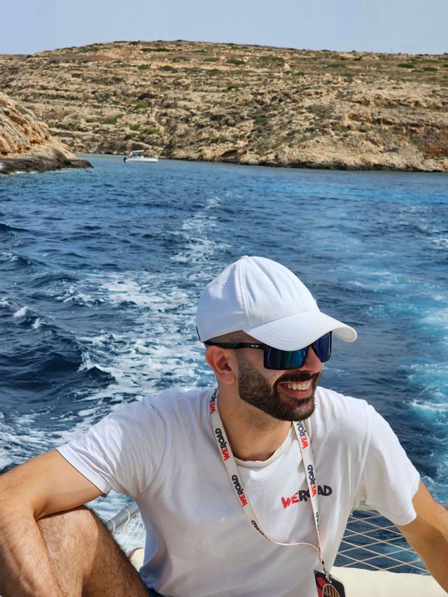 Un uomo sorridente con un cappellino bianco e una maglietta WeRoad siede su una barca in movimento, con acqua blu e una costa rocciosa dietro di lui.