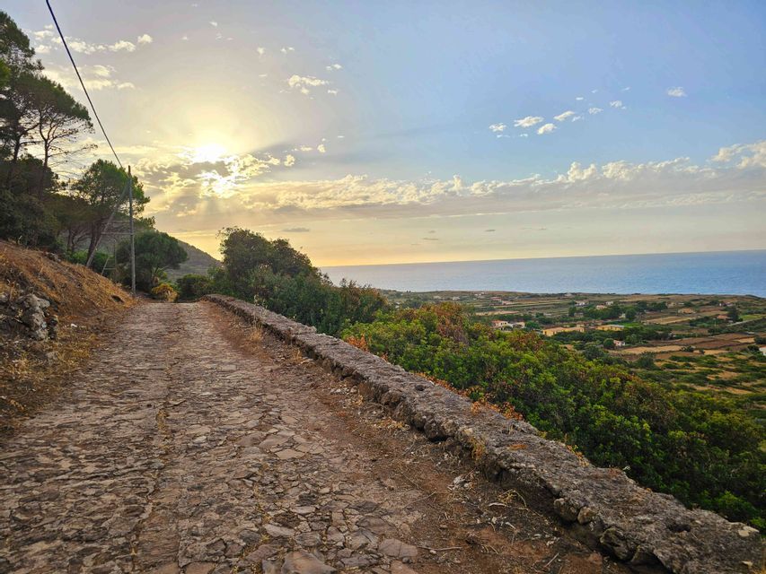 Un sentiero acciottolato si snoda lungo una collina che domina una verde pianura costiera e il mare al tramonto.