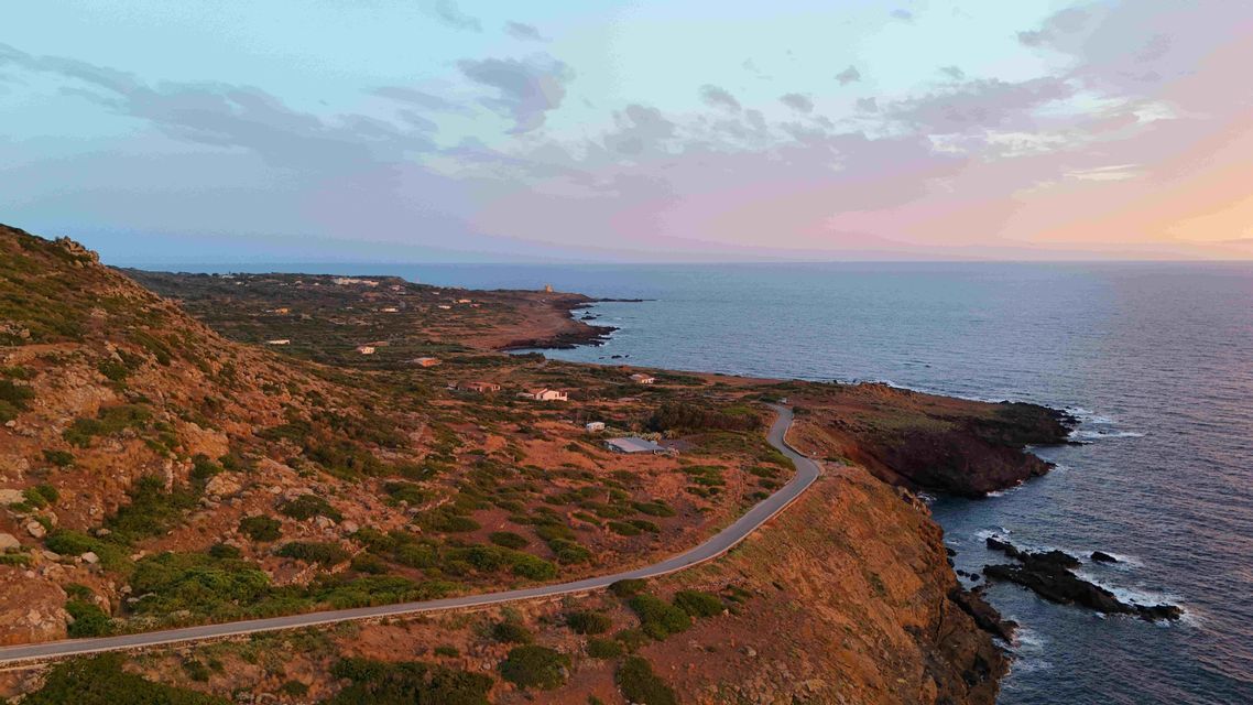 Veduta aerea di una strada tortuosa lungo una costa aspra e collinare al tramonto, sul mare.