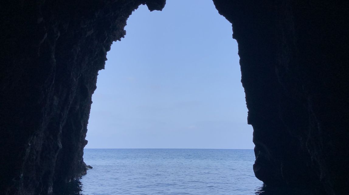 Vista dall'interno di una buia grotta rocciosa che si affaccia su un mare blu calmo sotto un cielo sereno.