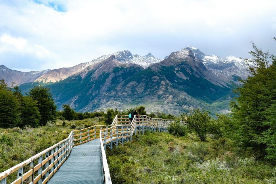 Una passerella in metallo e legno si snoda attraverso una valle verde verso una catena di grandi montagne innevate sotto un cielo parzialmente nuvoloso.