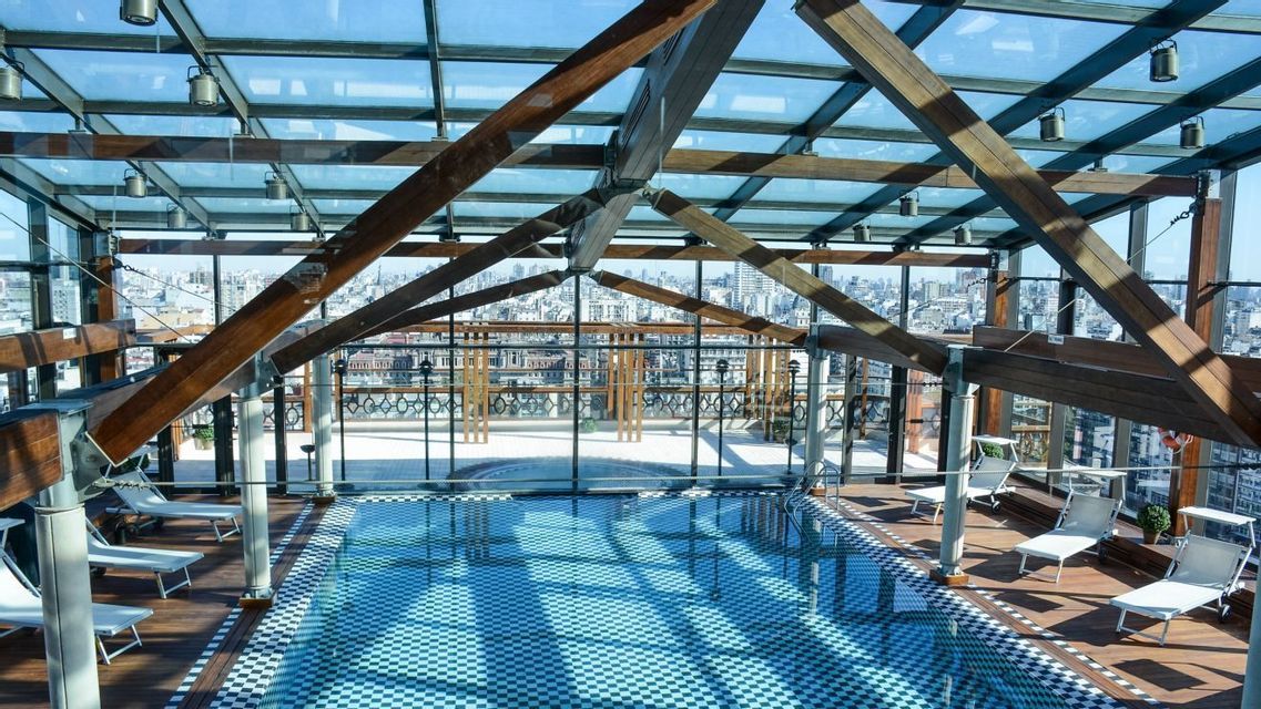 Una piscina coperta sul tetto con piastrelle a scacchiera e sedie a sdraio, circondata da pareti in vetro che si affacciano sullo skyline cittadino.