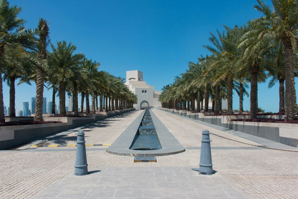 Un'ampia passerella pavimentata, fiancheggiata da palme e con un elemento d'acqua centrale, conduce a un edificio moderno, con lo skyline di una città sullo sfondo.
