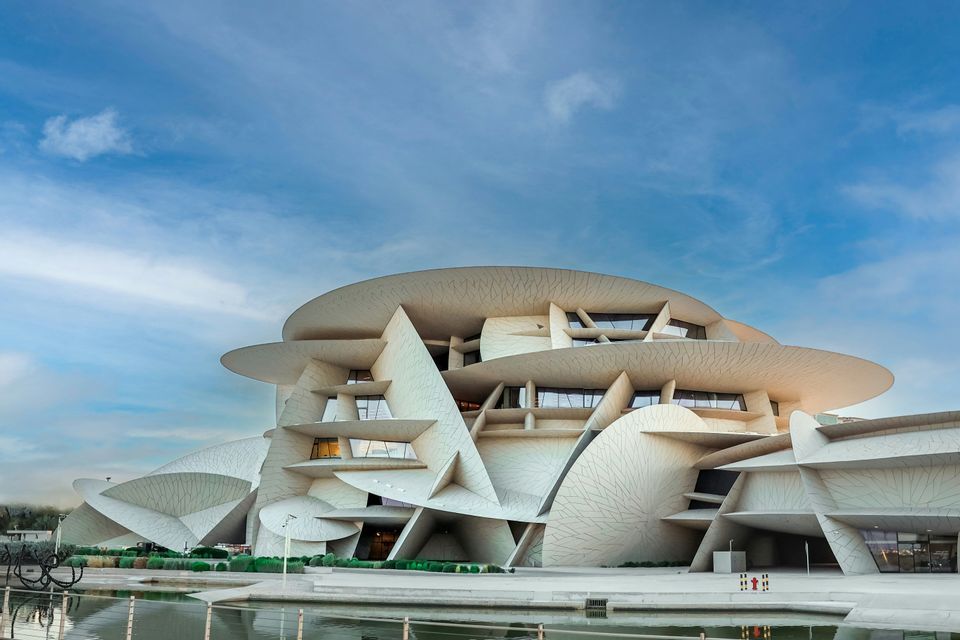 Un edificio moderno con strutture a dischi intrecciati si erge accanto a uno specchio d'acqua sotto un cielo blu.